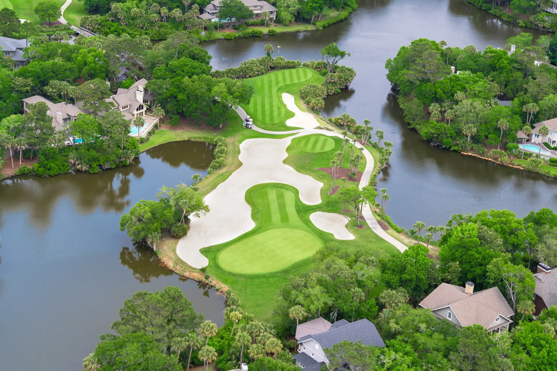 Golf hole surrounded by sand bunkers and water with nearby houses.