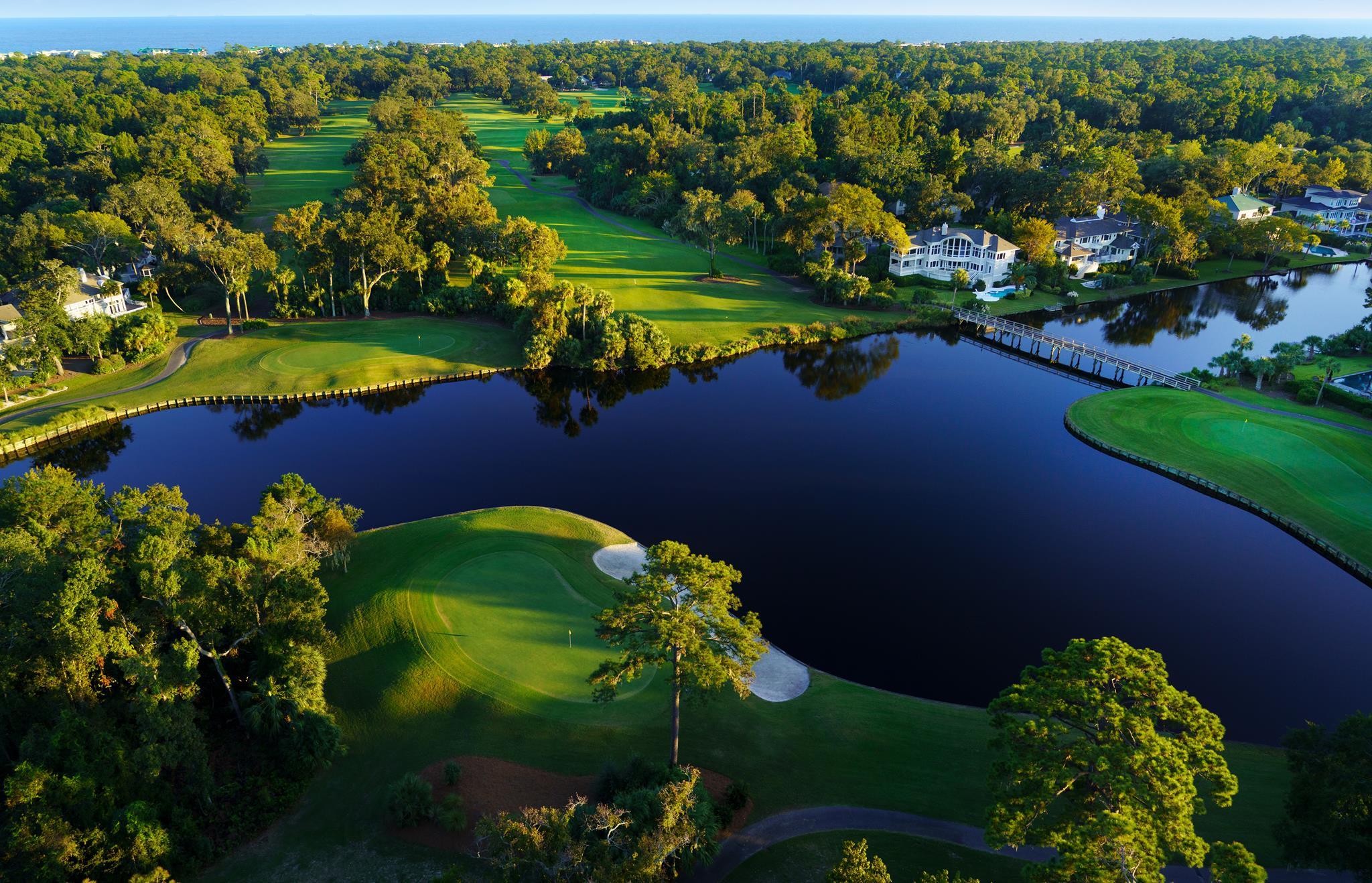 A scenic aerial view of a golf course with lush fairways surrounded by water and nearby houses, creating a serene coastal setting.