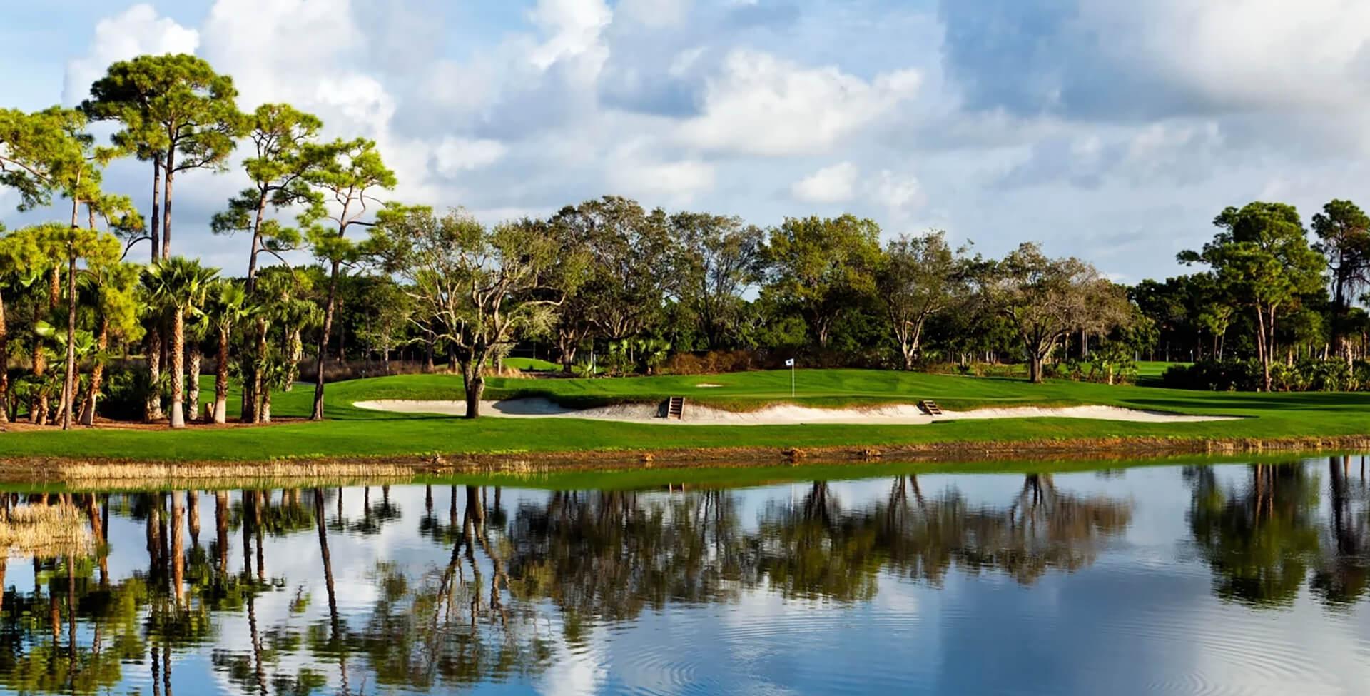 A green with white sand bunkers near a calm pond, surrounded by palm and oak trees with reflections visible in the water.