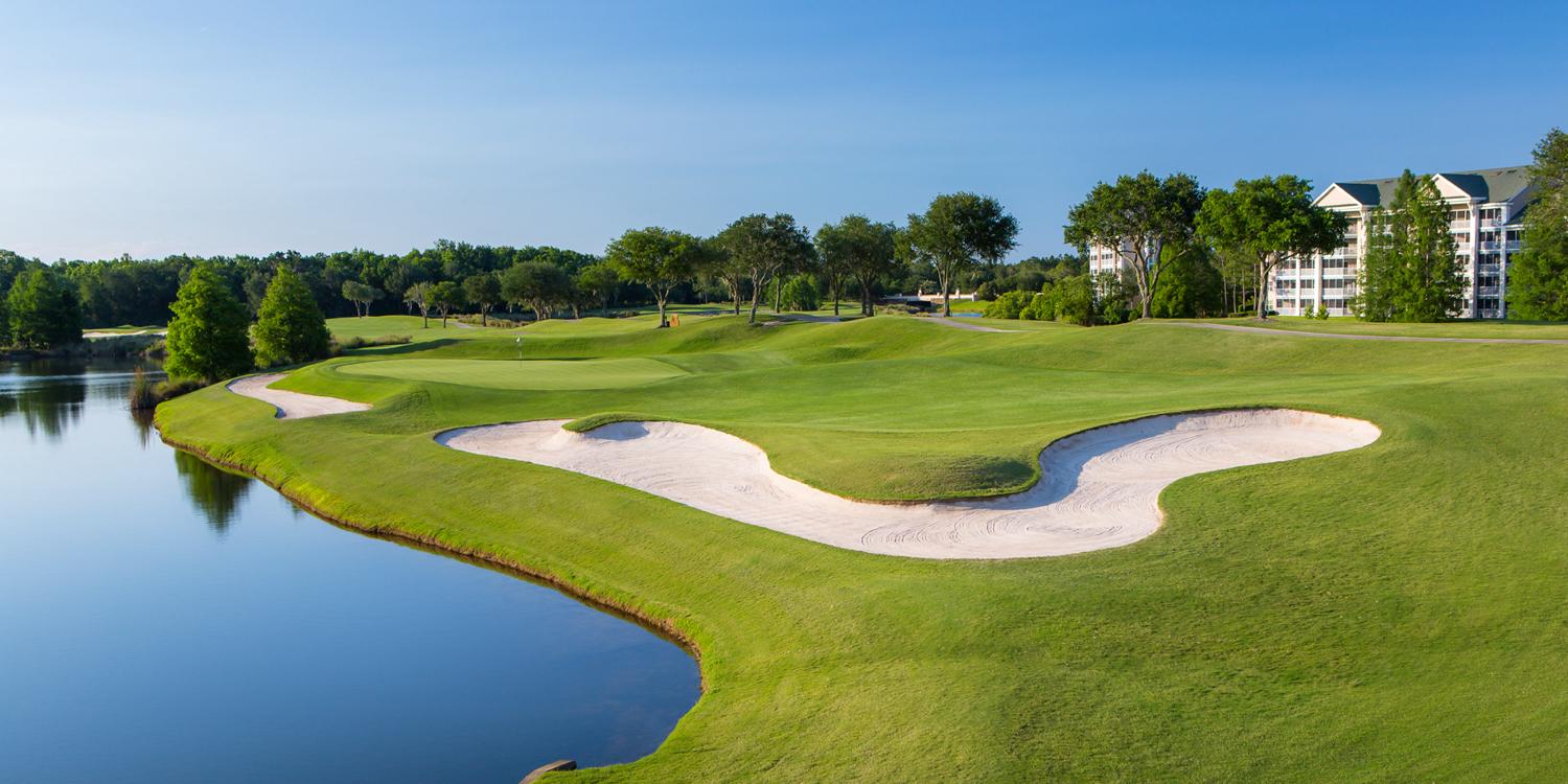 A scenic golf hole features pristine bunkers alongside a peaceful lake with modern buildings in the background.