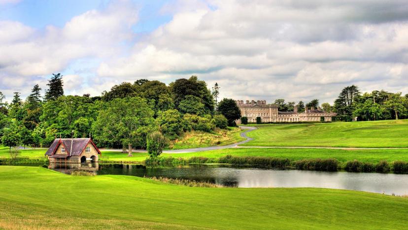 Rolling fairways lead to a green near a tranquil lake and historic building.