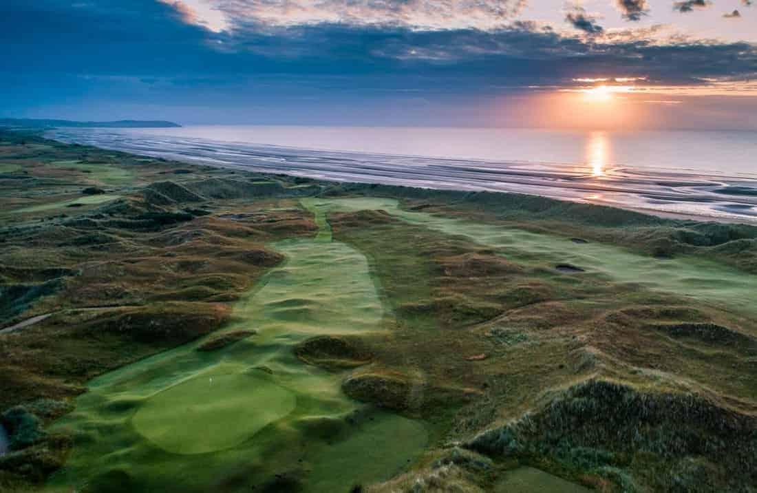A stunning aerial view of a links course with the sun rising over the ocean.