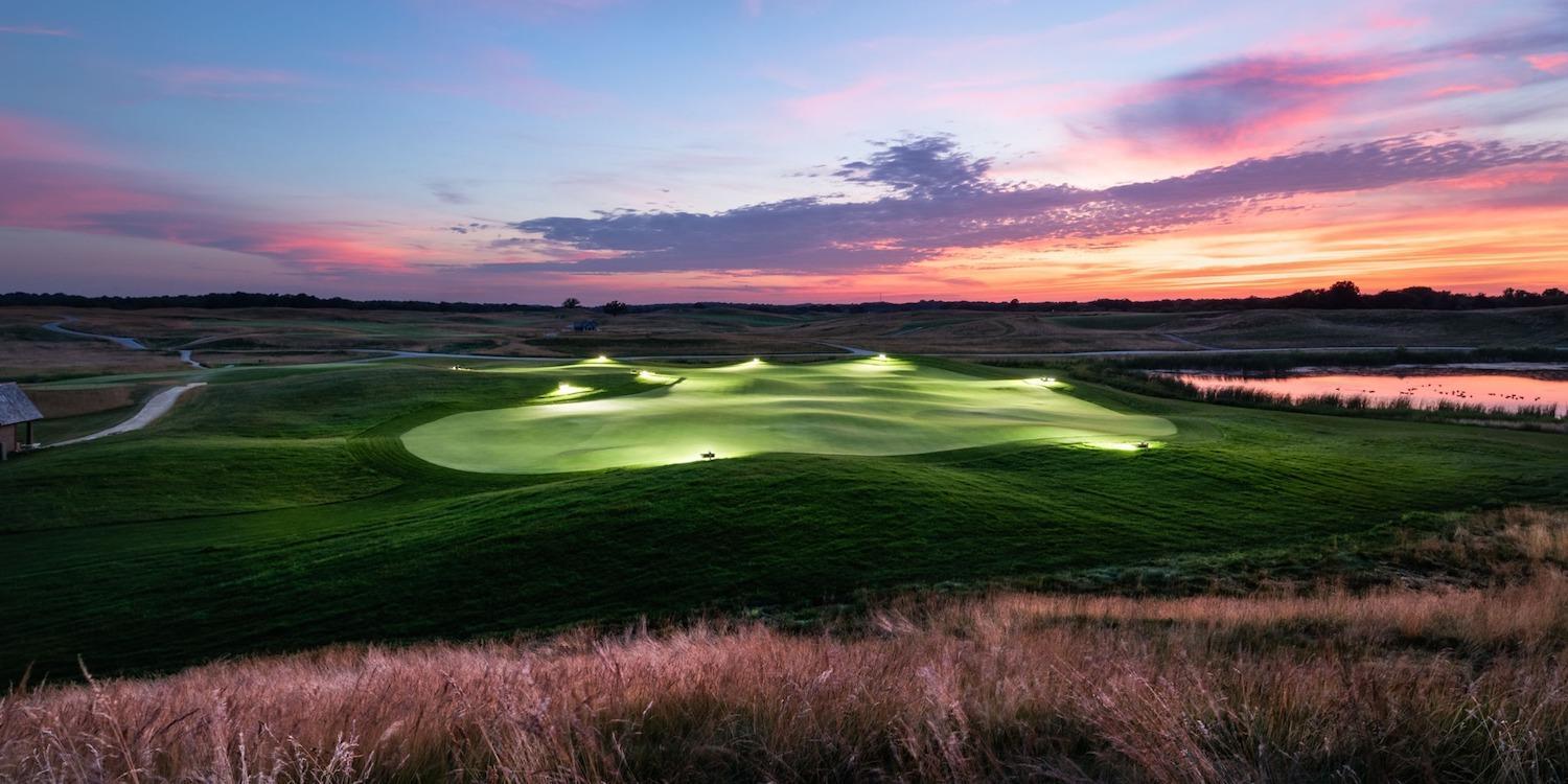 Panoramic view of a green being lit up by flood lights under a purple evening sky at sunset