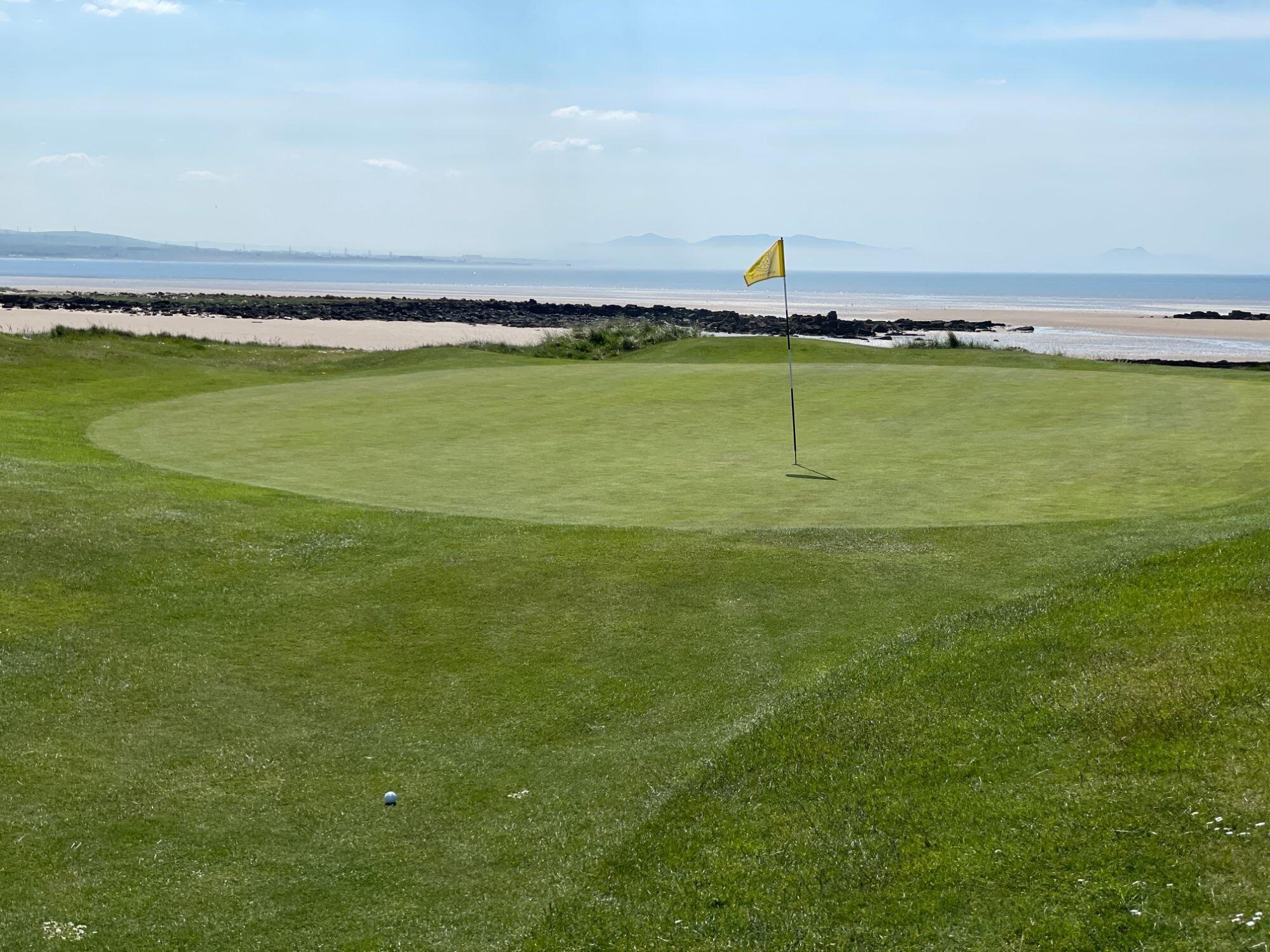 A scenic seaside green with a yellow flag overlooking sandy beaches and distant hills.