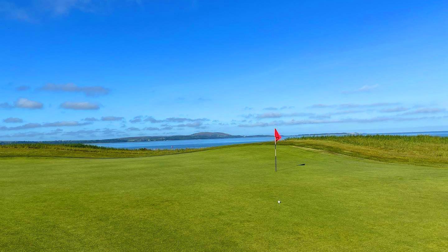 A pristine golf green with a flagstick, overlooking a calm body of water under a clear blue sky.