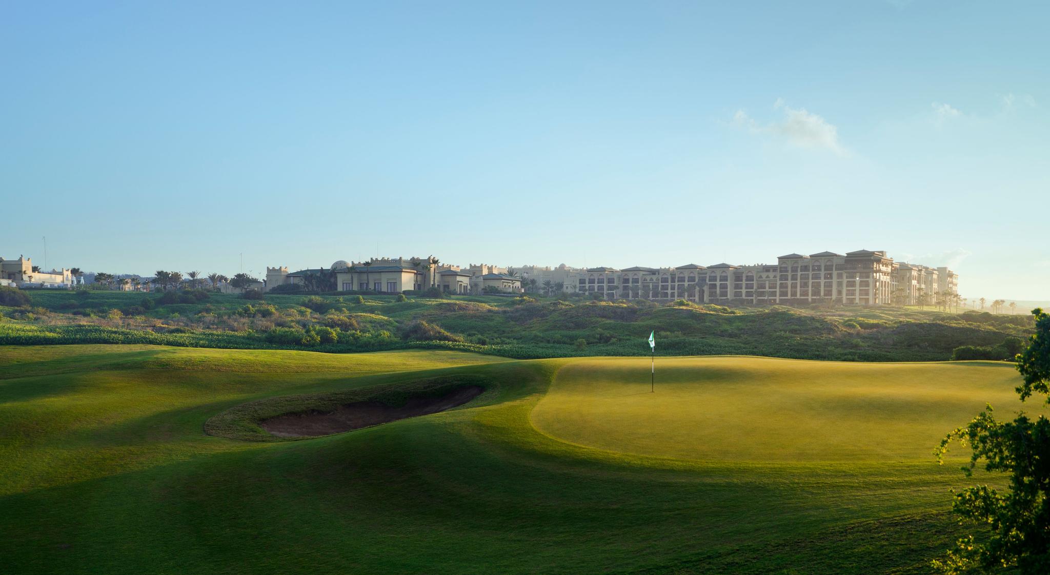 A smooth green next to a sand bunker with the Mazagan Beach & Golf Resort building in the back