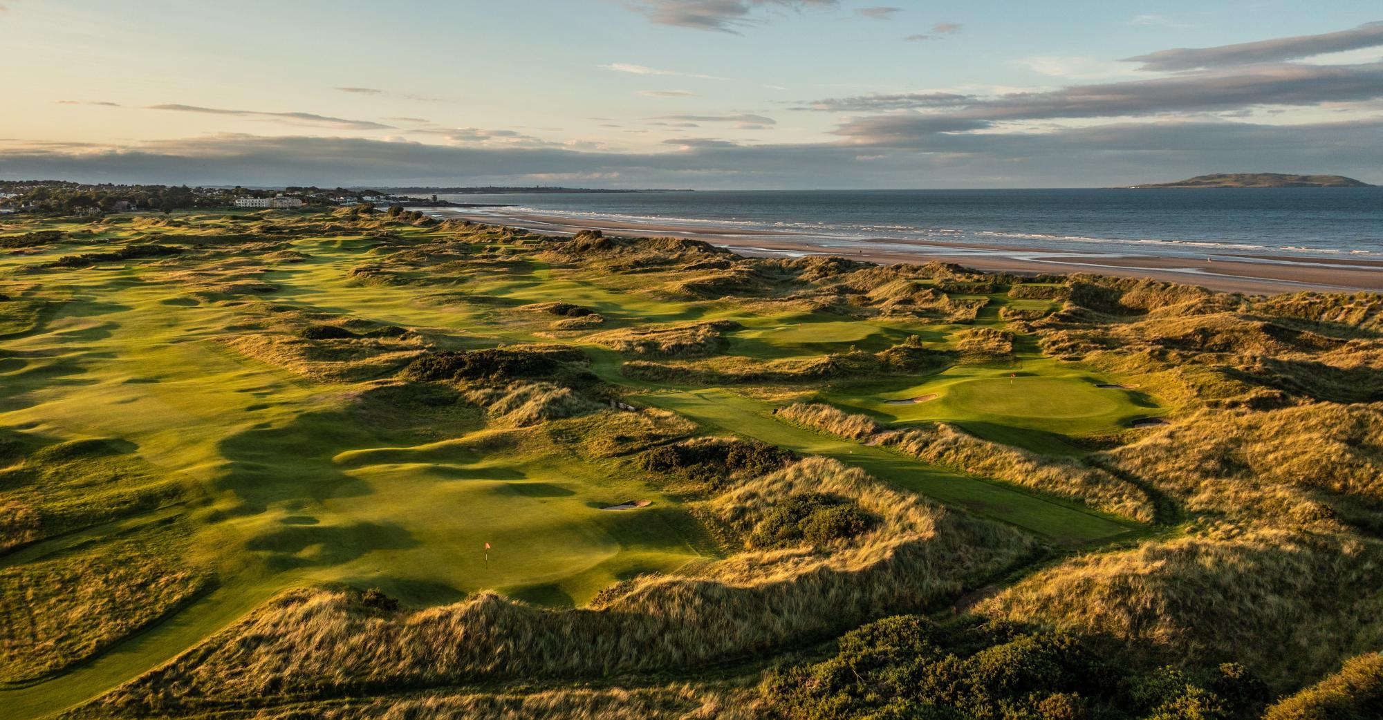 Rolling green dunes of a golf course run alongside the ocean at sunset.