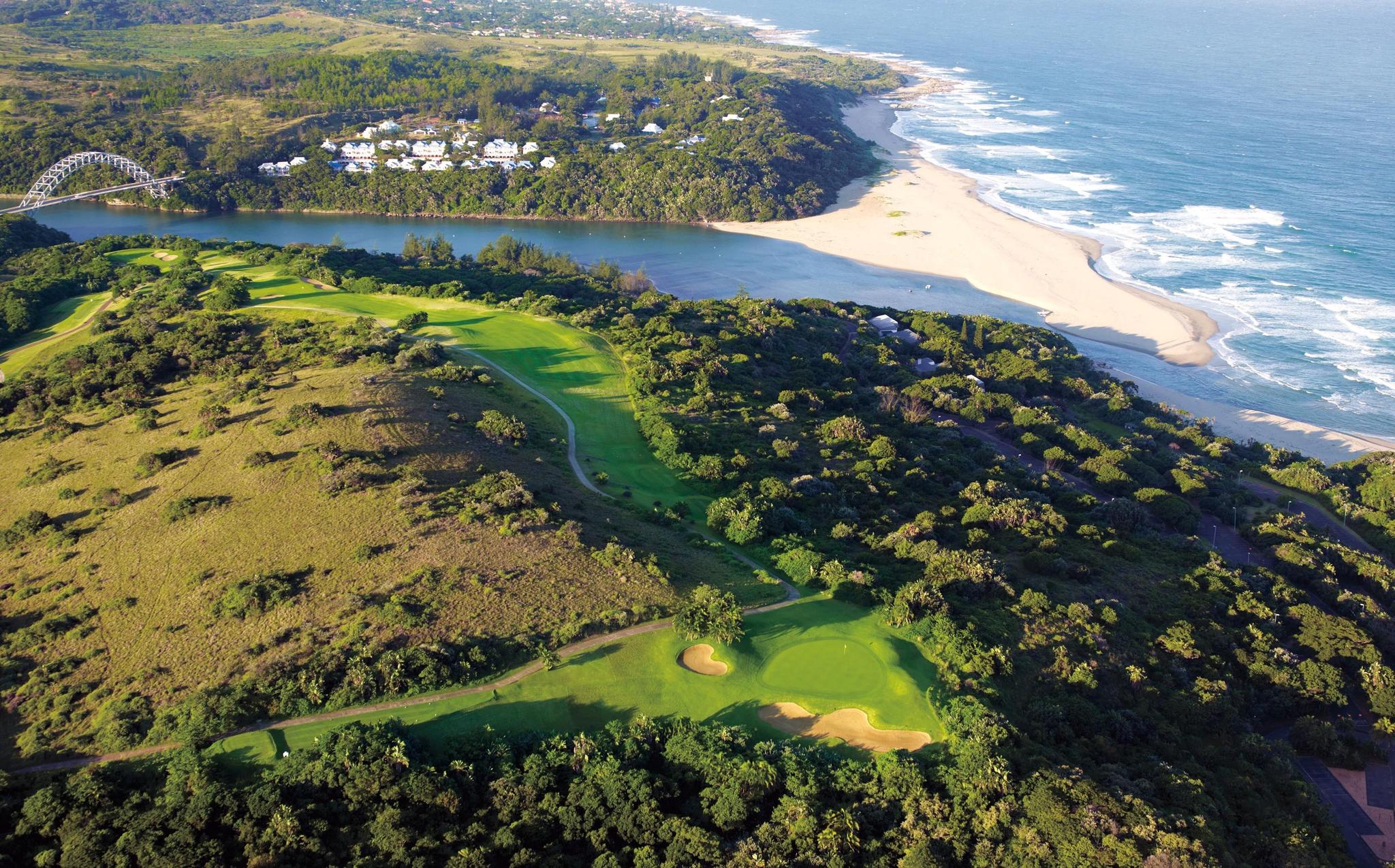 A coastal golf course nestled between hills, a river, and the ocean with a bridge in the distance.