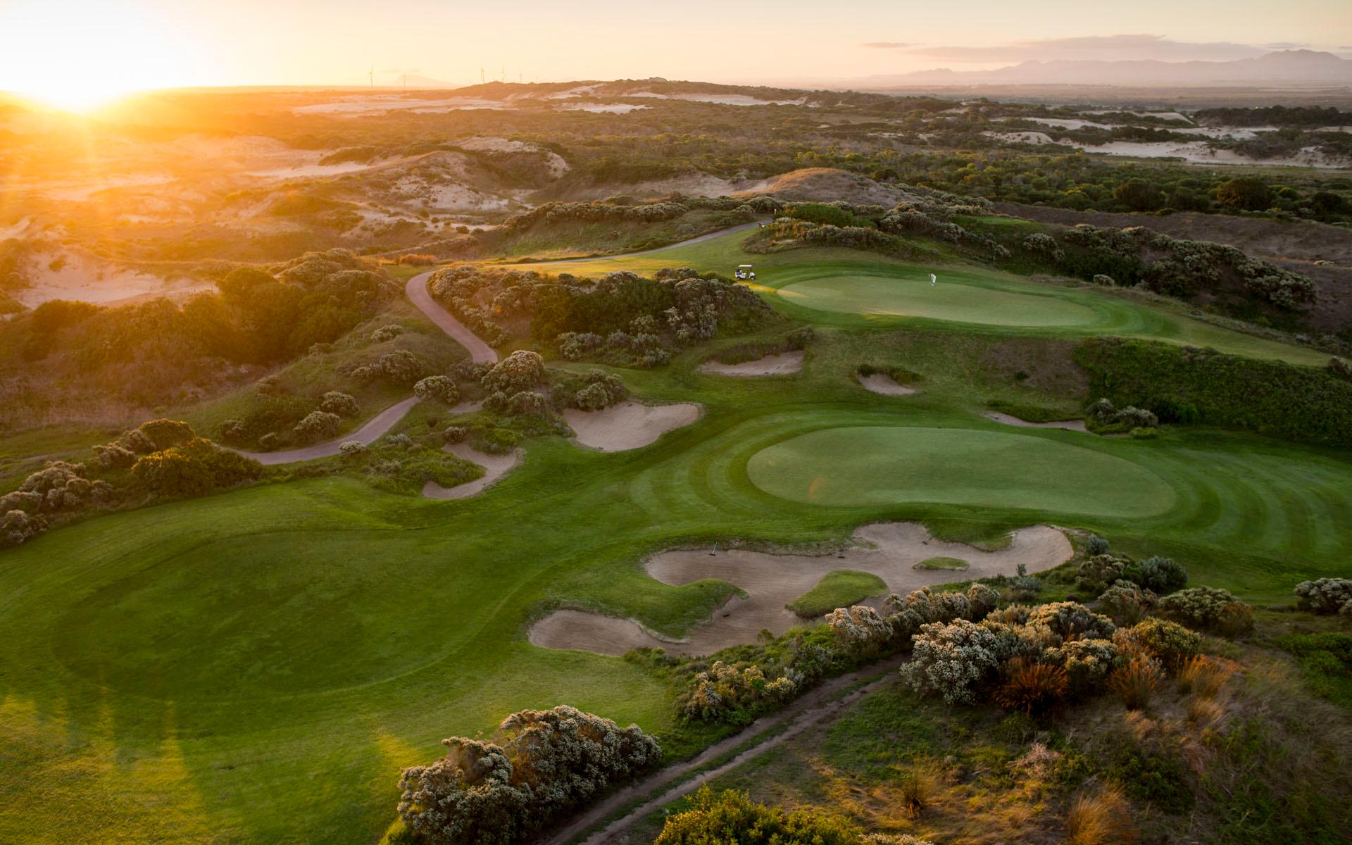 A dramatic sunset view over a rugged golf course with winding paths and sand traps.