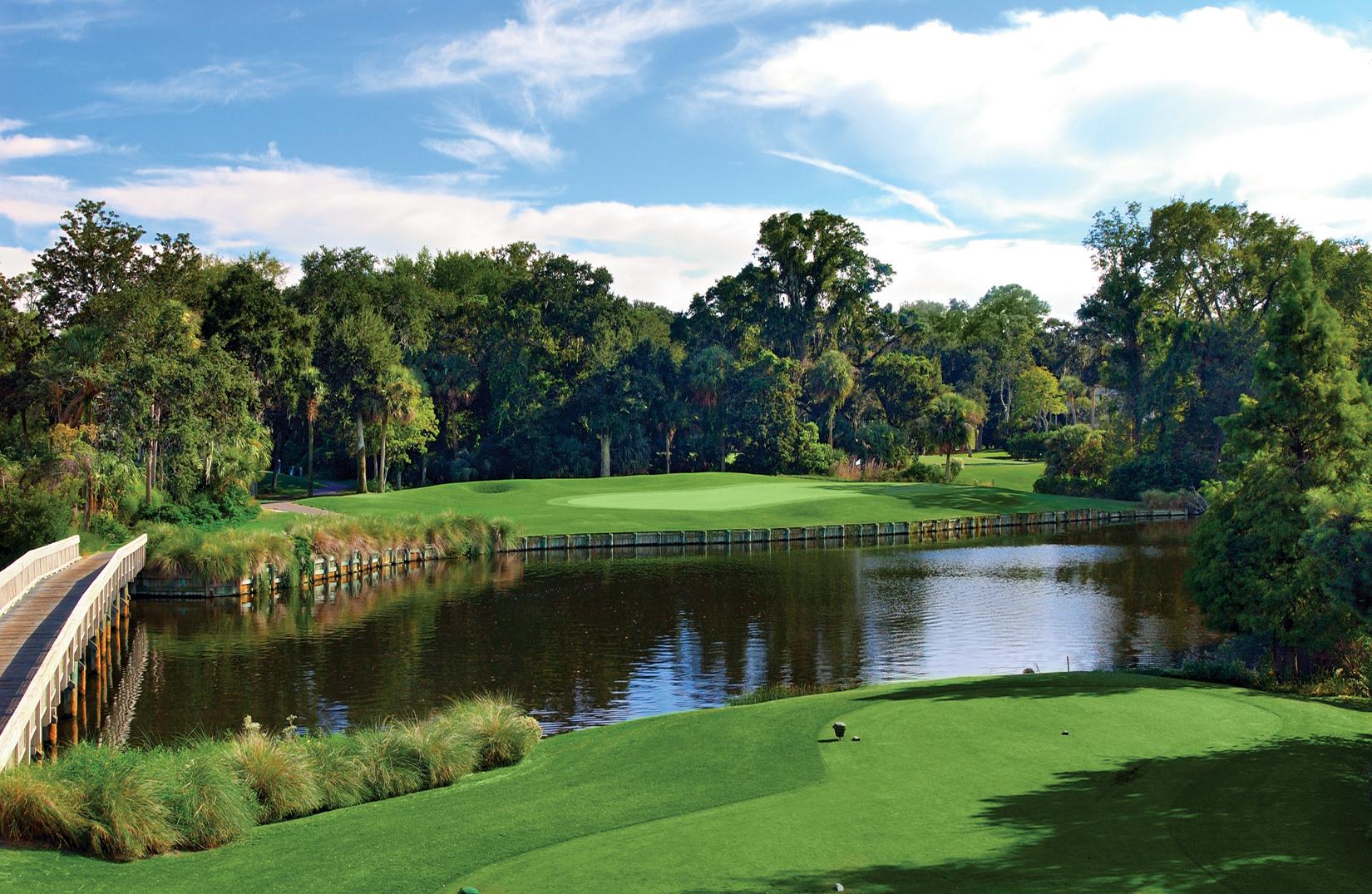 A beautiful water-surrounded golf hole with a wooden bridge connecting the fairways amidst dense greenery.