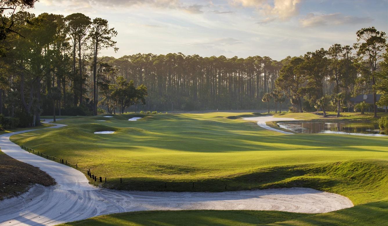 A rolling green fairway with sand bunkers and a winding path through a forested backdrop.