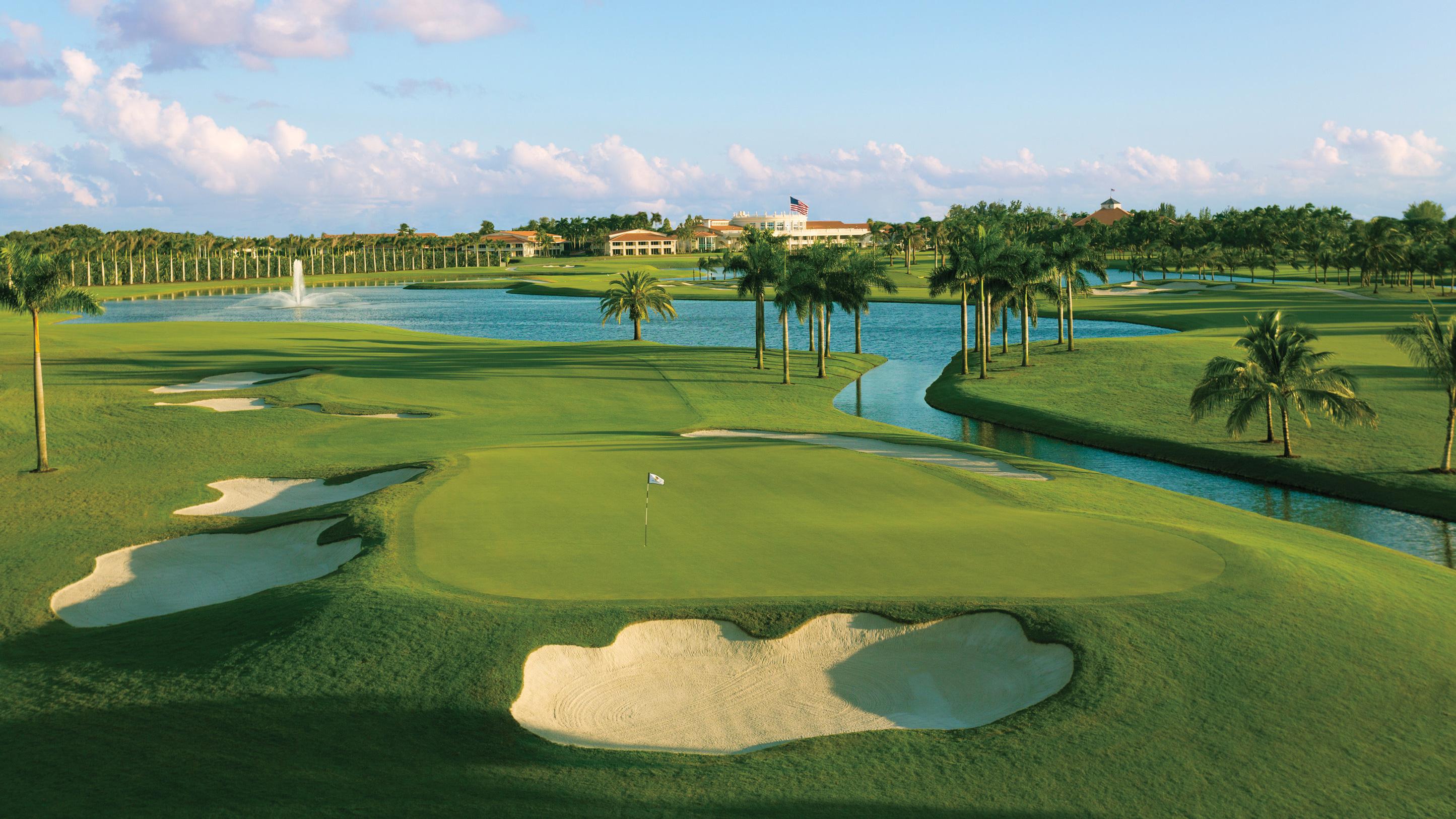 Elevated manicured green surrounded by sand bunkers and palm trees with the clubhouse in the distance