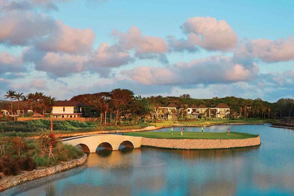 Golfers play on an island green connected by a stone bridge, surrounded by water.