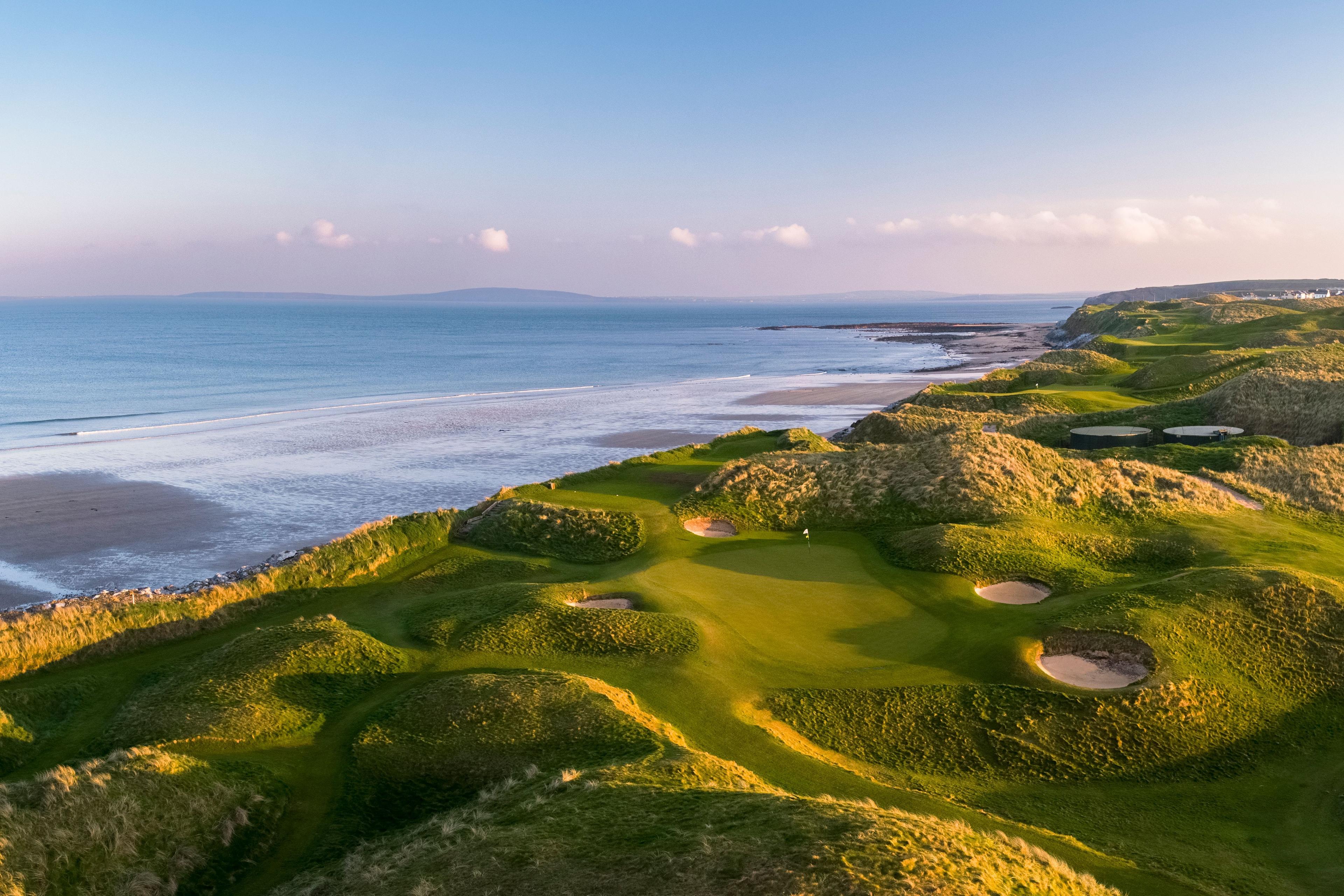 A beautifully framed green surrounded by dunes, overlooking the ocean under a pastel sky.