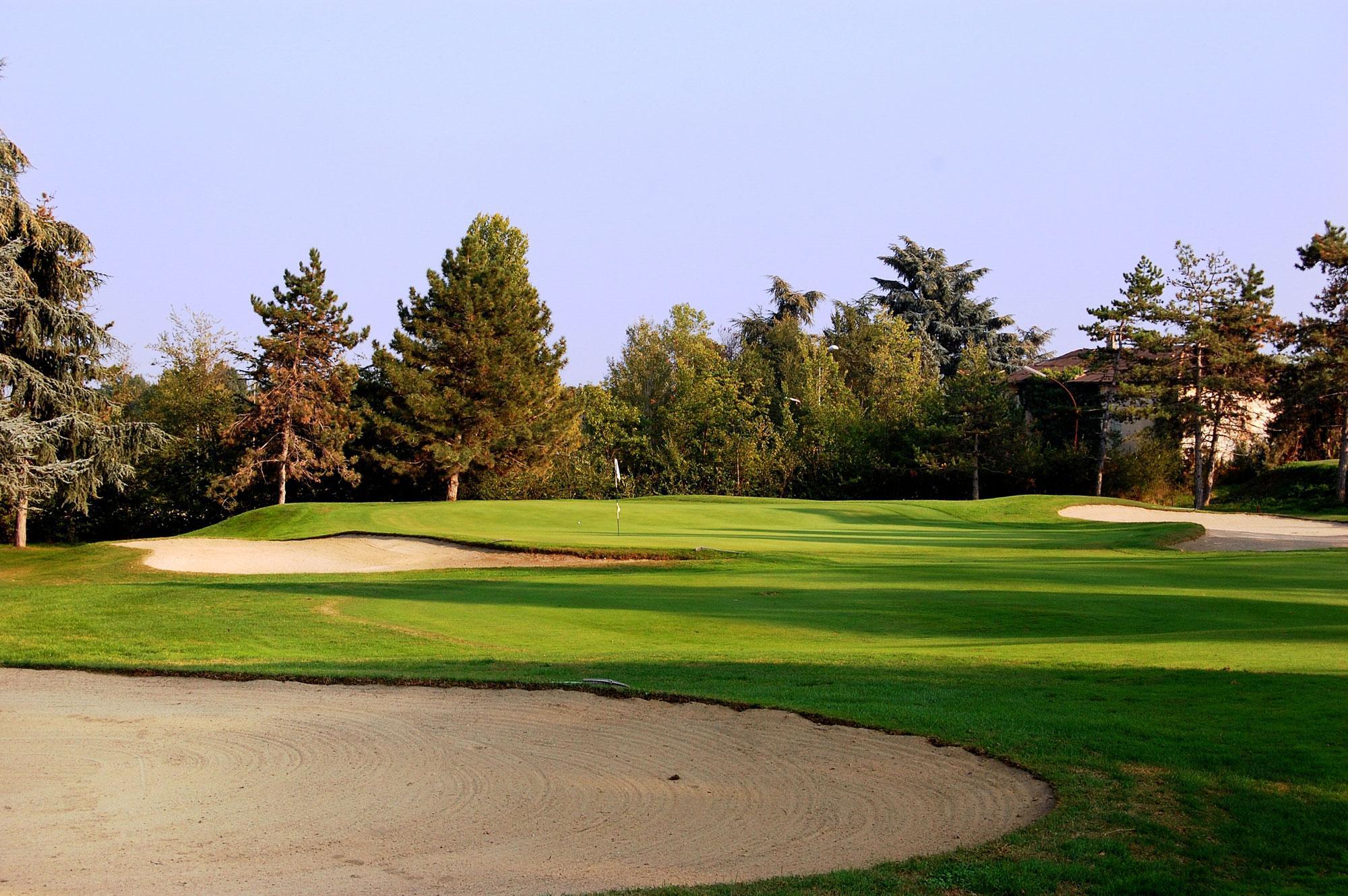 A golf green bordered by sand bunkers and surrounded by lush trees under a clear sky.