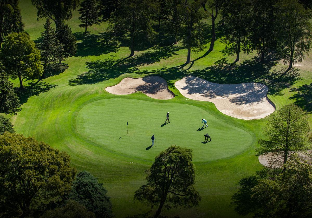An aerial view of golfers playing on a green surrounded by sand bunkers and trees