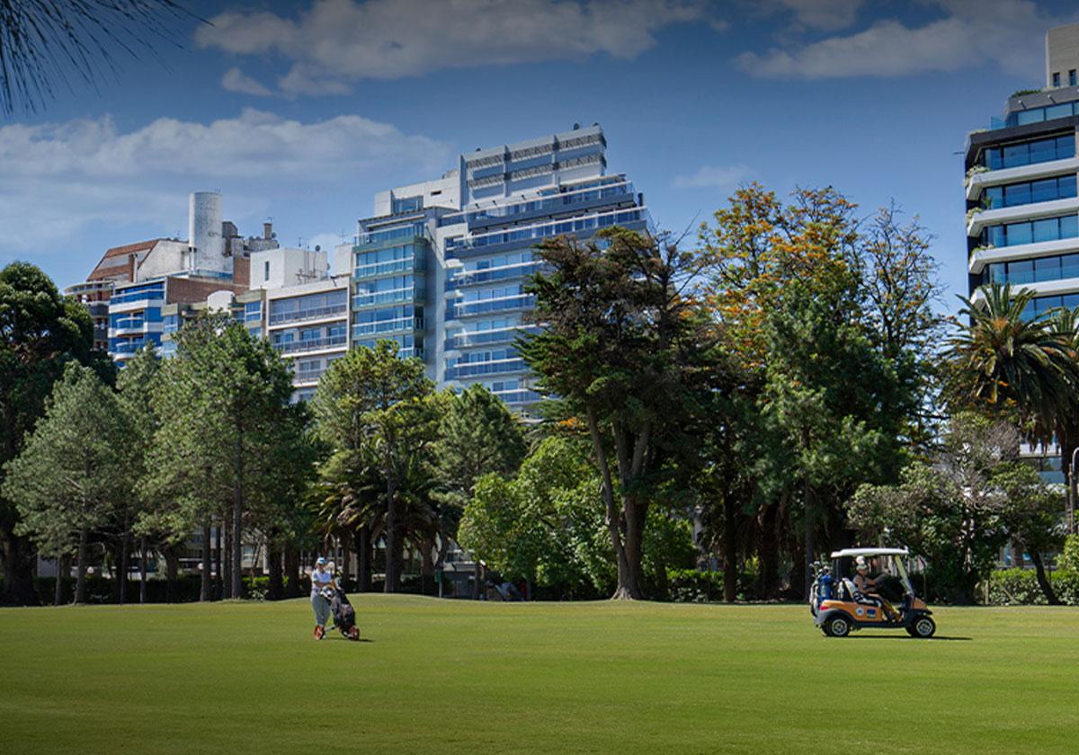 A golf course with players and a golf cart set against a backdrop of city buildings