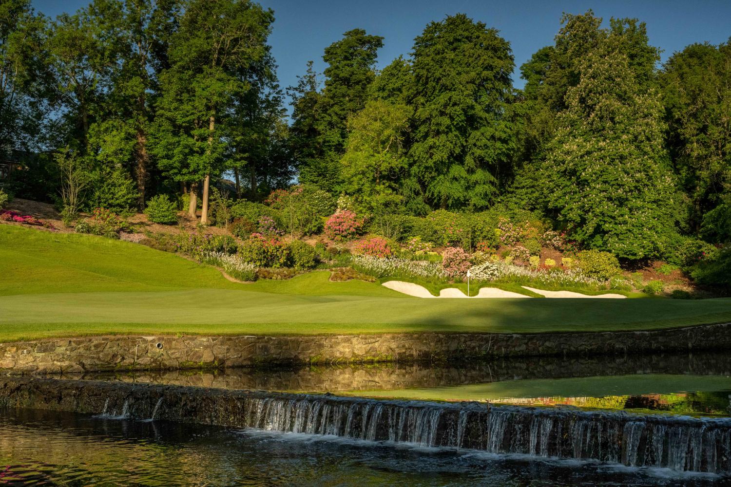 A picturesque waterfall flows beside a pristine green, surrounded by colorful flowers.