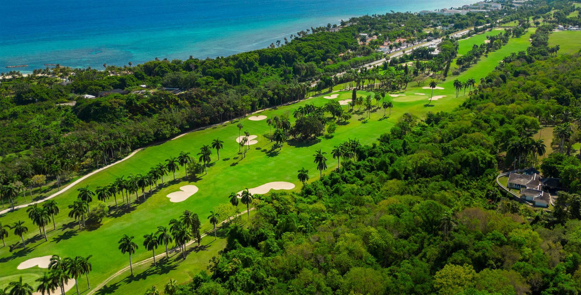 Aerial view of a coastal golf course lined with palm trees and turquoise waters.