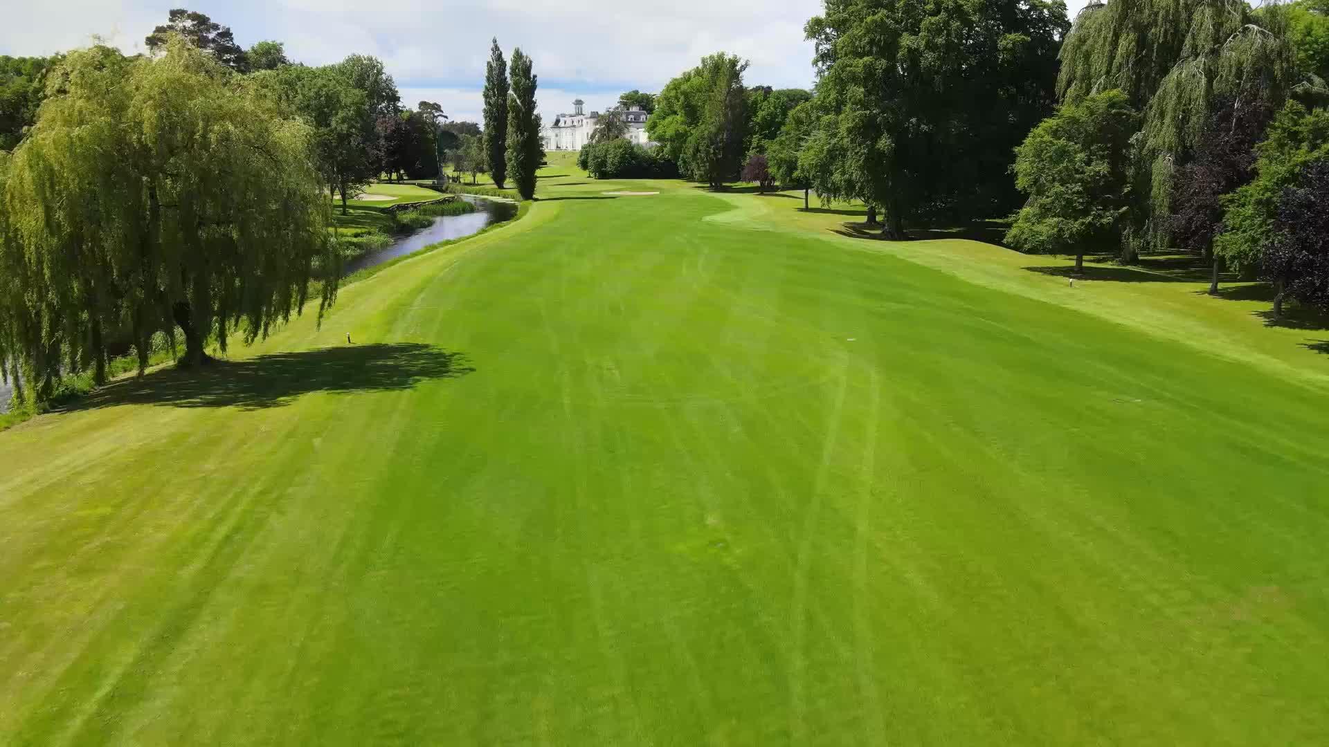 A long, tree-lined fairway with a meandering river leading towards a grand estate in the distance.