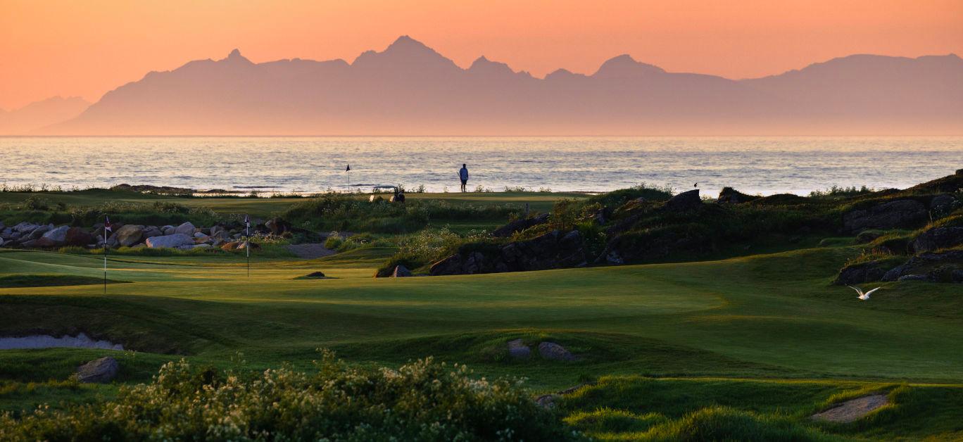 A golfer standing on an elevated green under evening skies with a mountain in the background