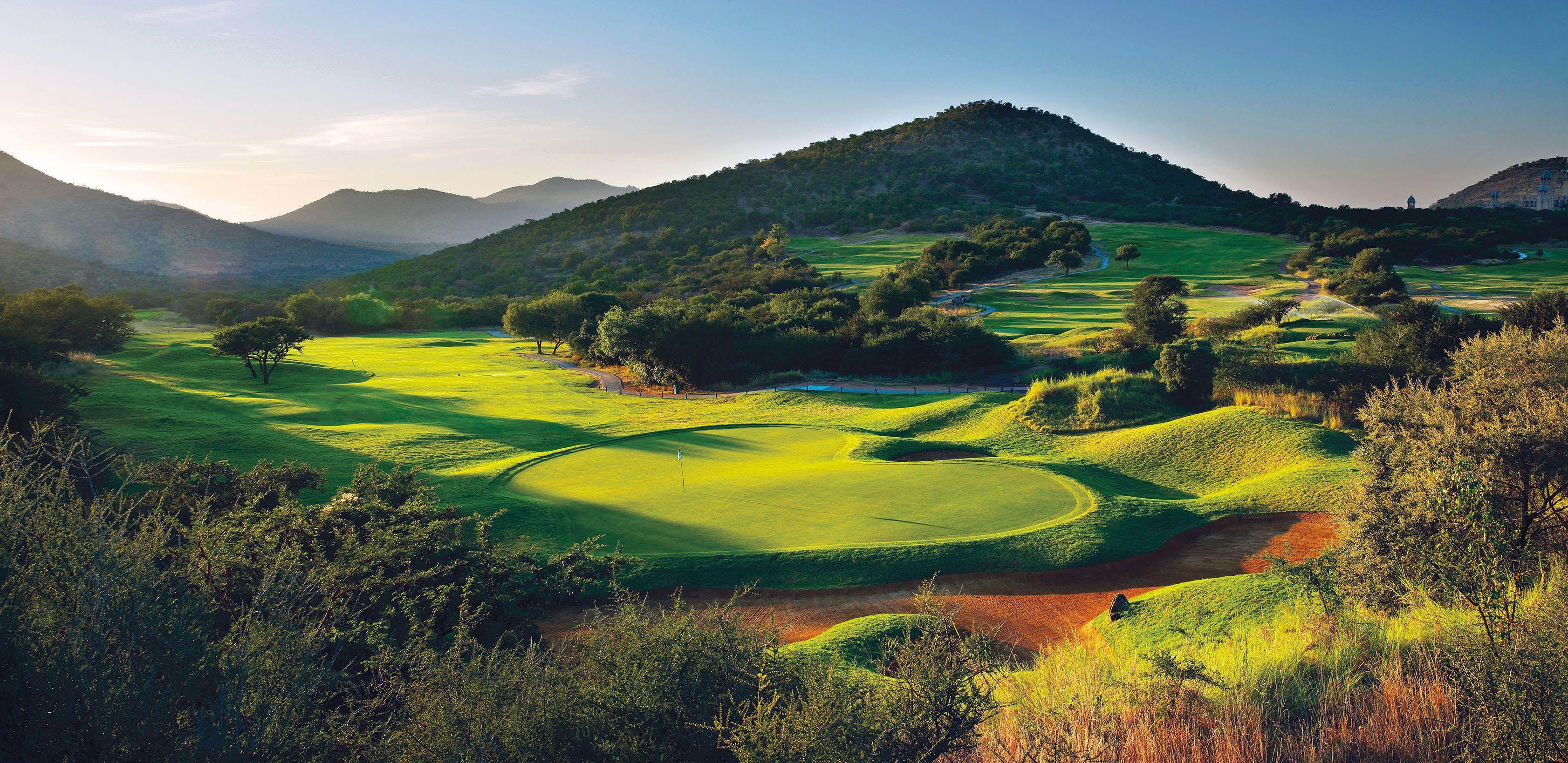Rolling green fairways winding through a mountainous landscape at sunrise.
