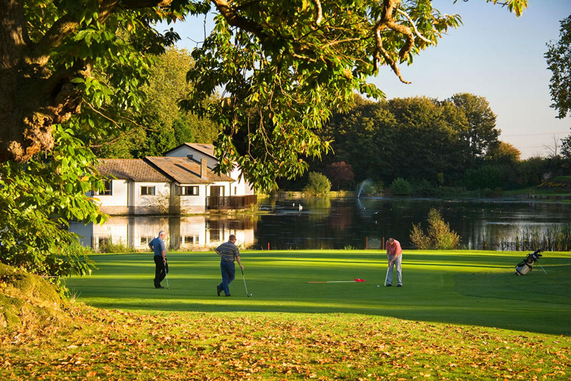 Golfers enjoy a game near a water hazard during late afternoon.