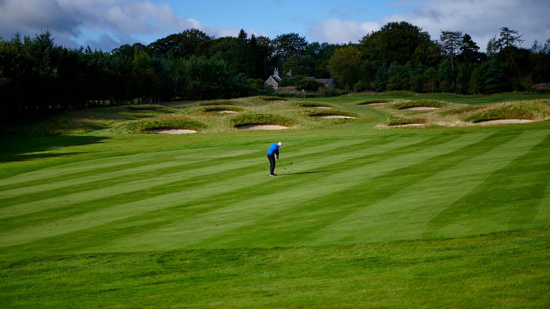 Golfer hitting towards the greens surrounded by deep bunkers with a tree line in the back