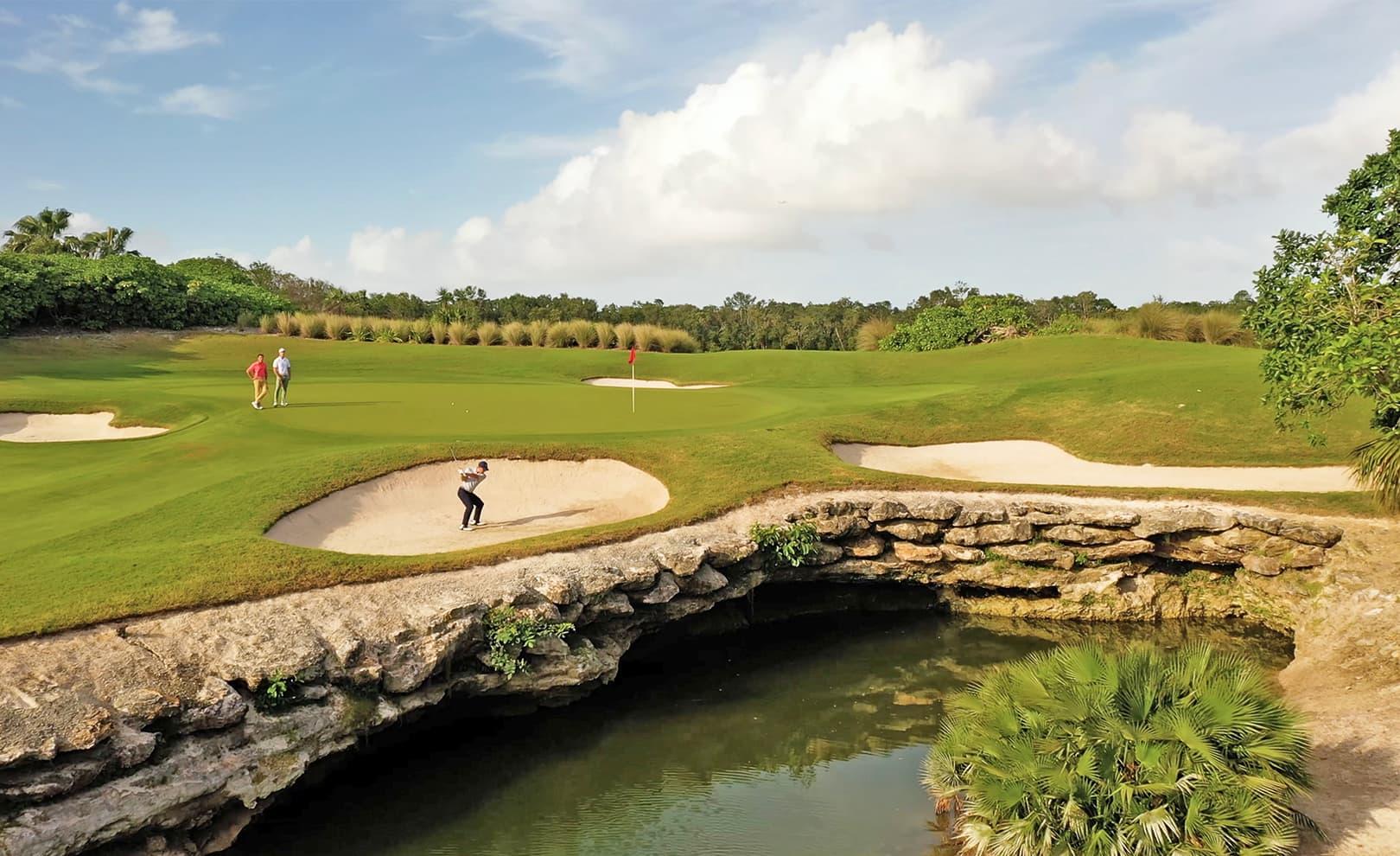 Golfer taking a shot from a bunker near a rocky ledge with clear water below.