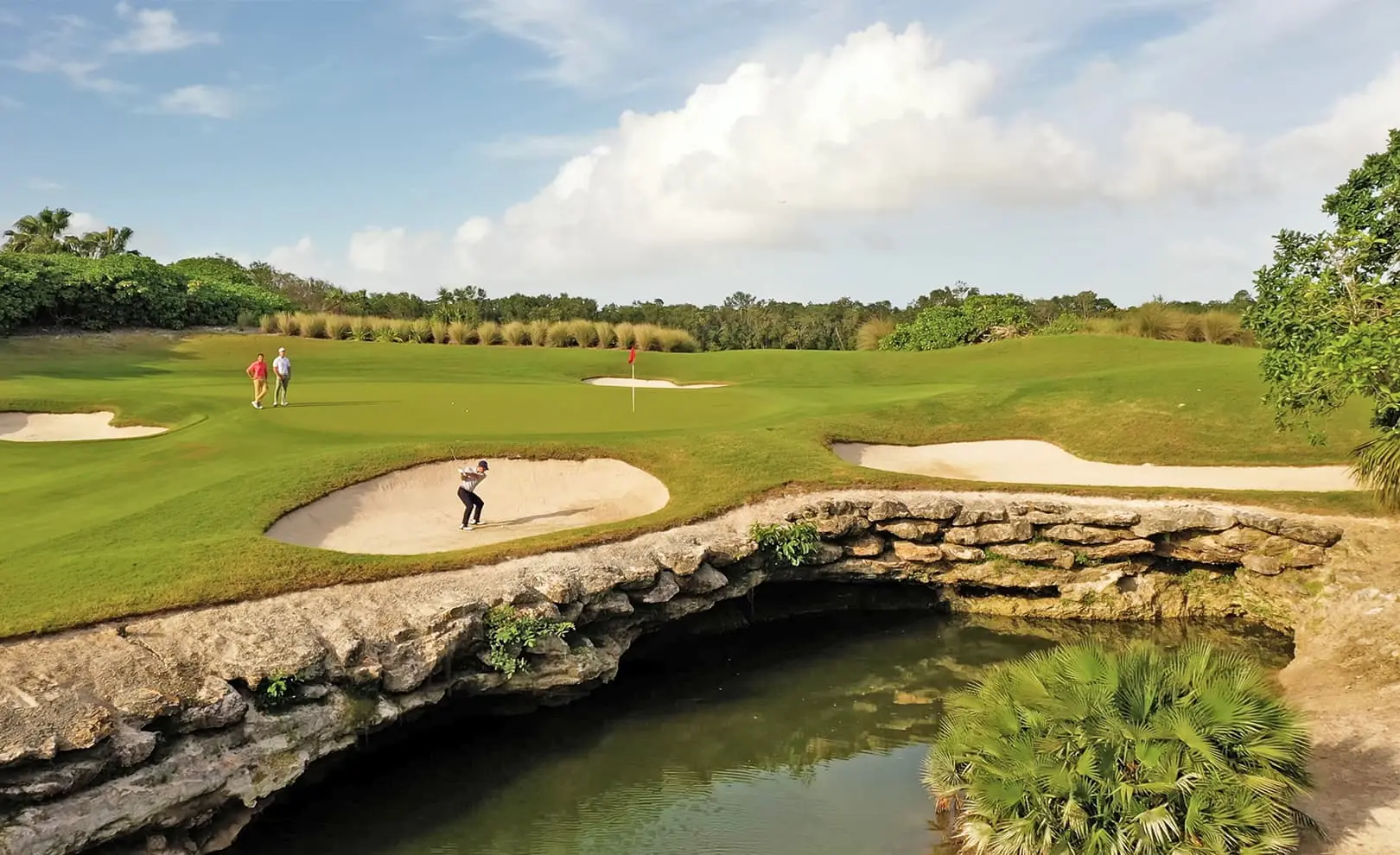 Golfer taking a shot from a bunker near a rocky ledge with clear water below.