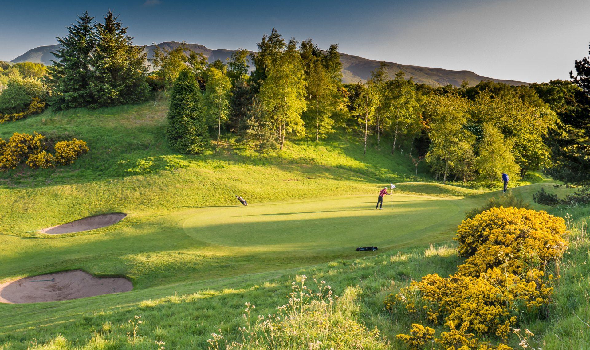 Two golfers play on a scenic green backed by hills and golden gorse bushes.