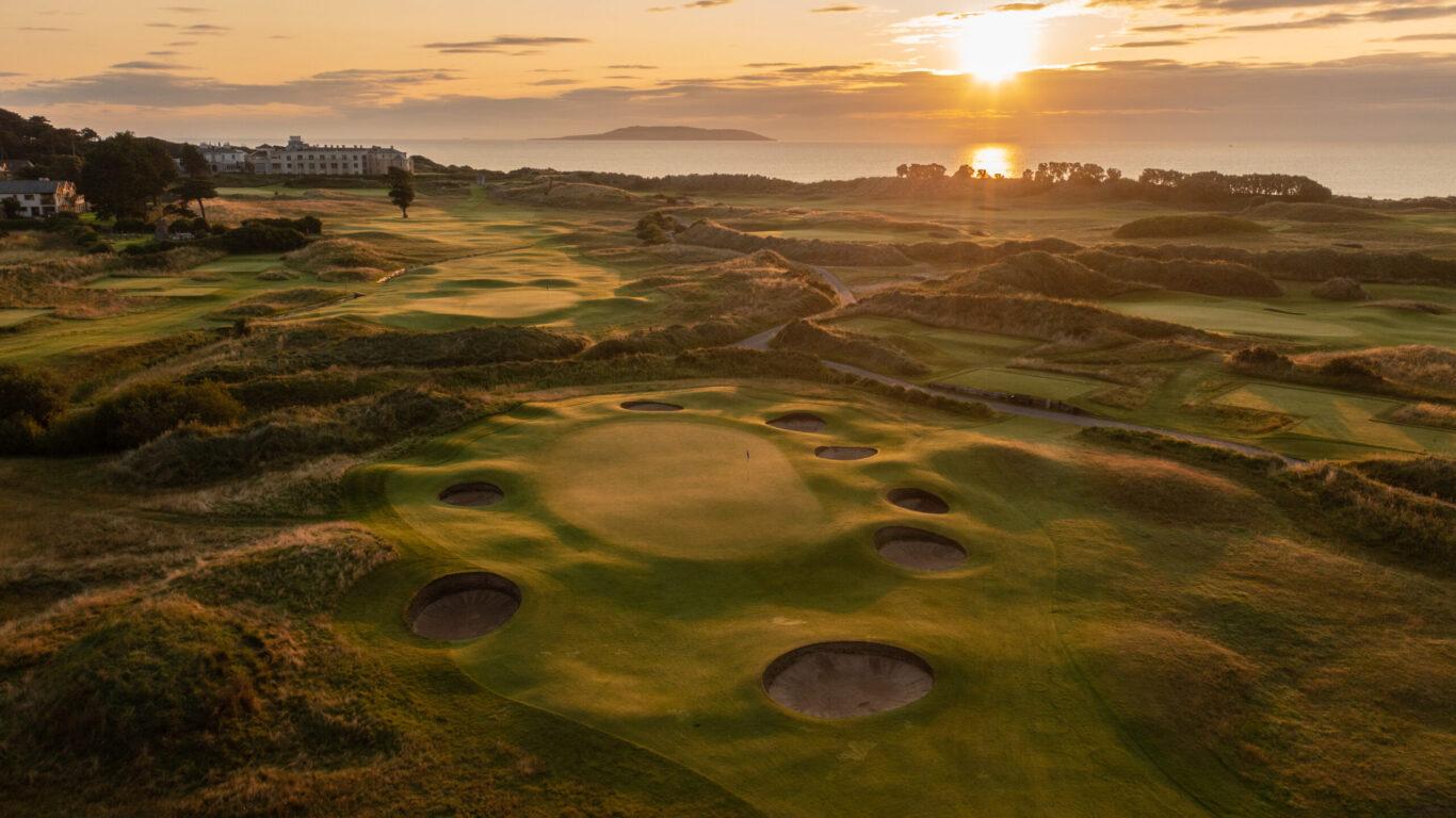 Sunset illuminates a dramatic golf green surrounded by circular bunkers near the sea.
