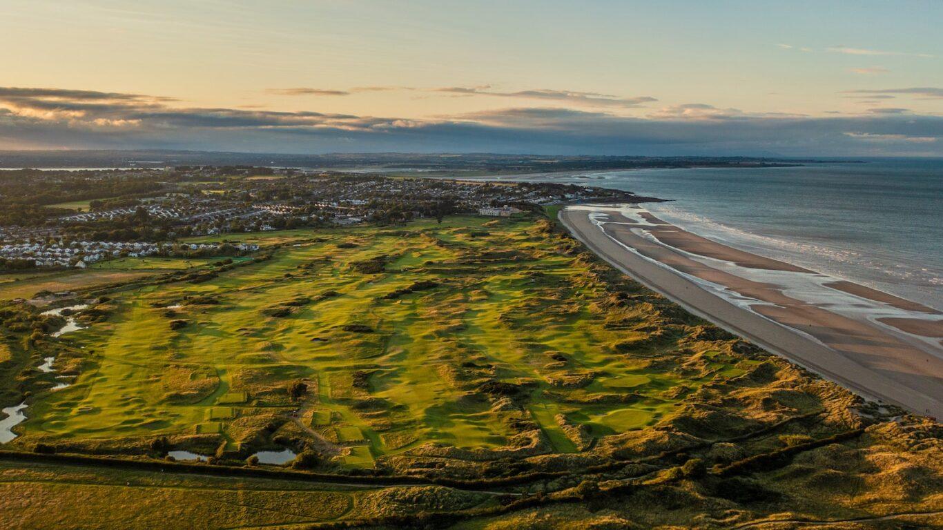 A wide view of a seaside golf course with nearby houses and sandy shores.