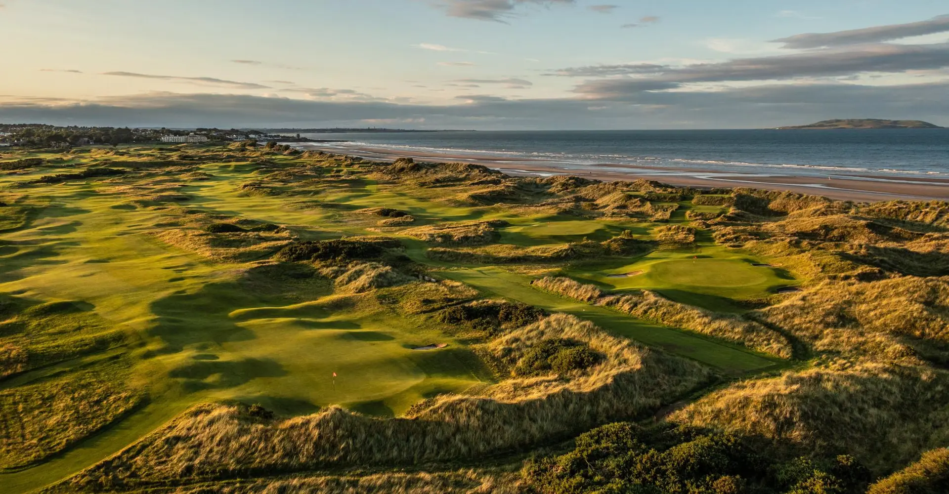 Rolling green dunes of a golf course run alongside the ocean at sunset.