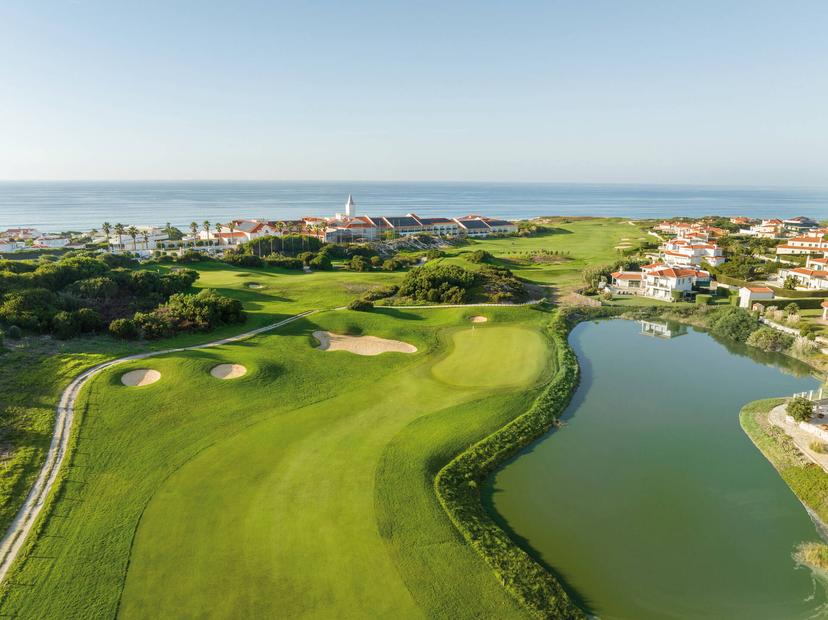 Aerial view of a fairway winding around a water hazard with the Marriott Praia d'El Rey in the background
