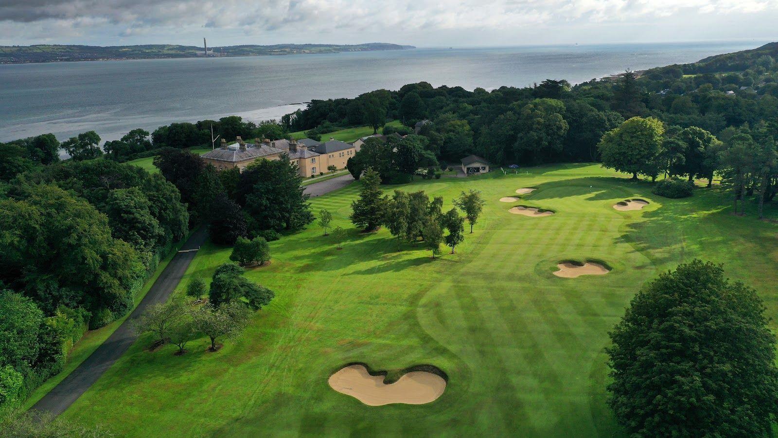 A historic clubhouse set beside tree-lined fairways with scenic sea views in the distance.
