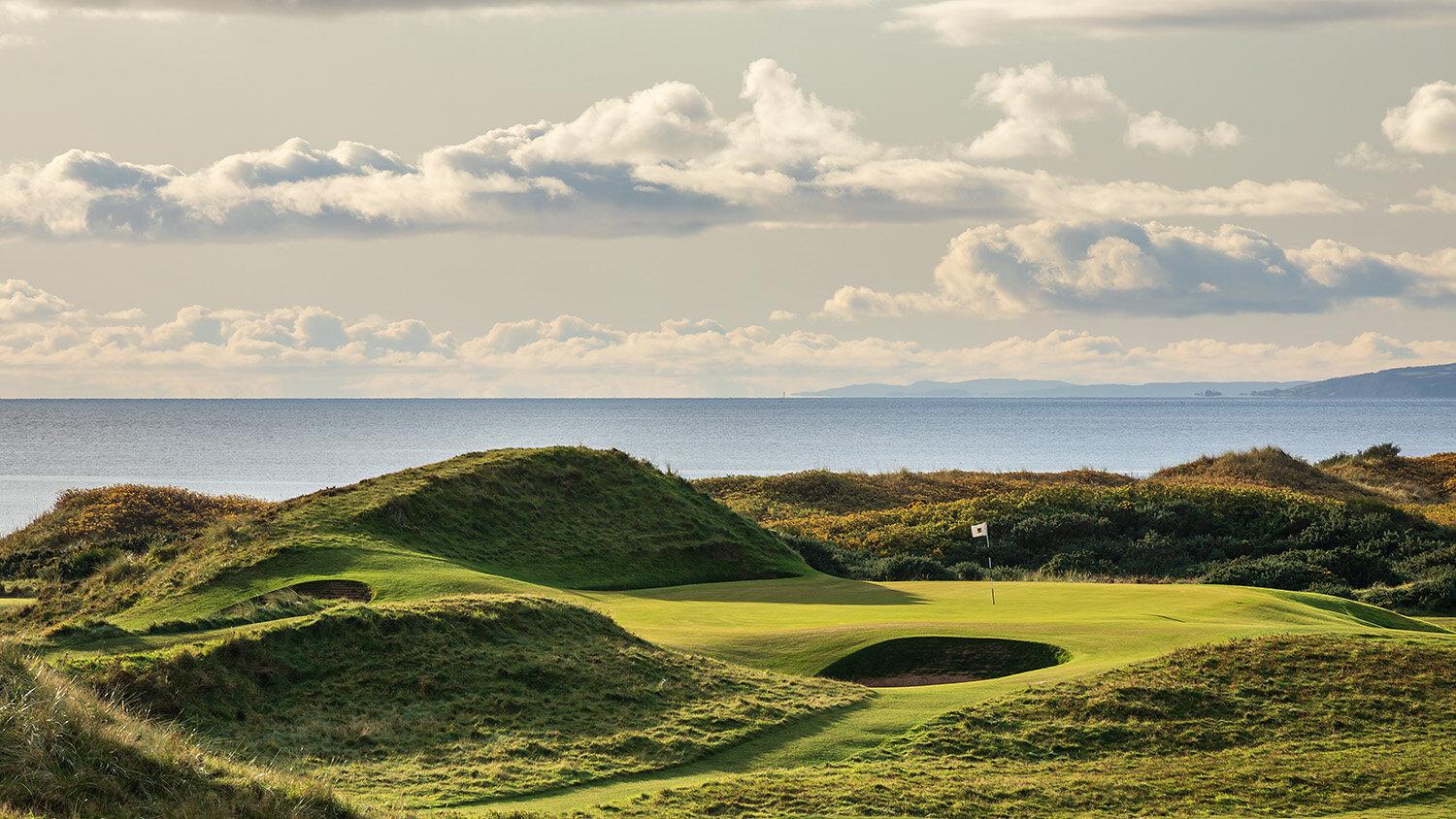 Hilly landscape around the well-kept green nestled with sand bunkers and ocean views in the distance