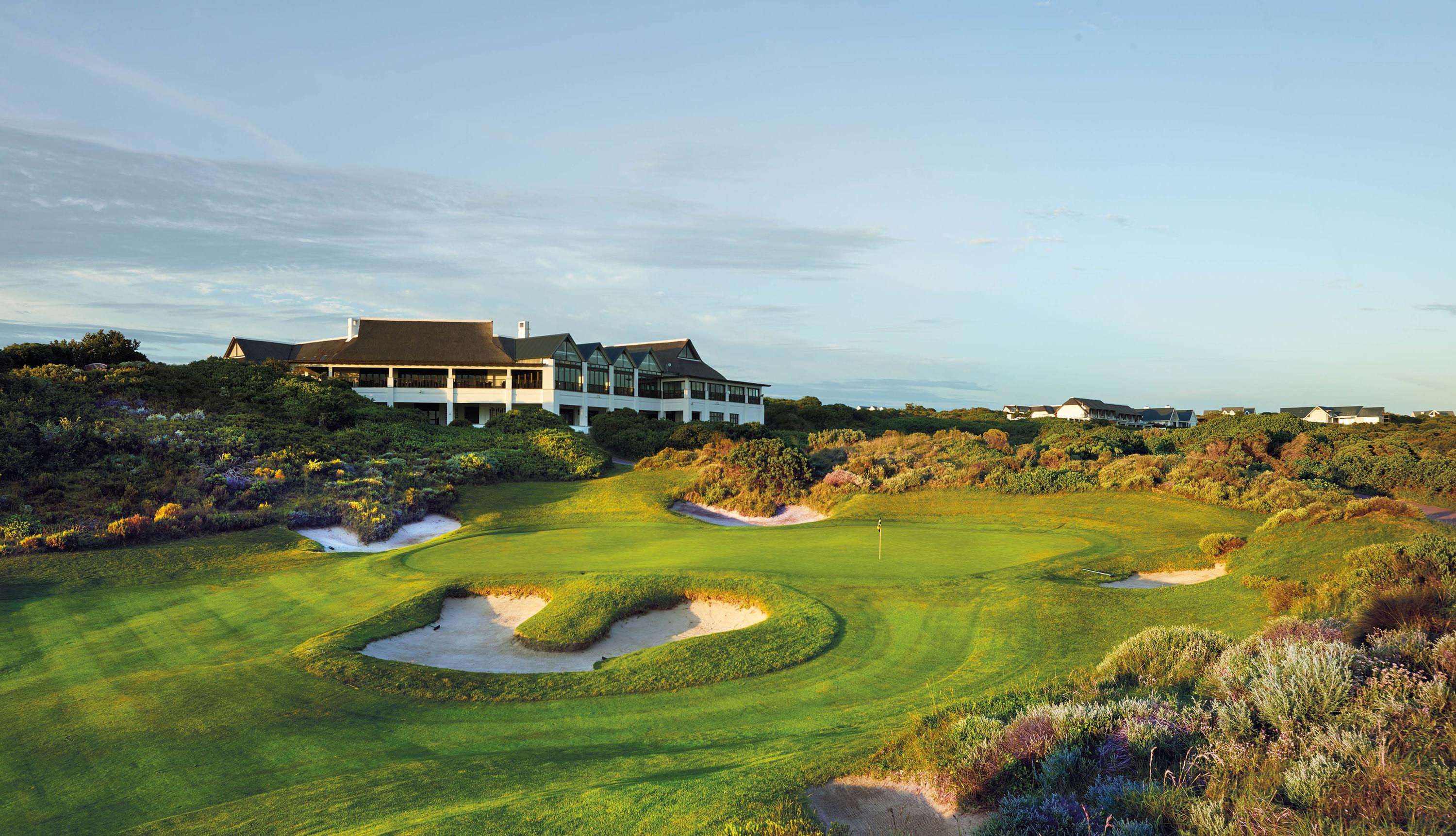 A golf green with deep bunkers, with a large modern clubhouse in the background.