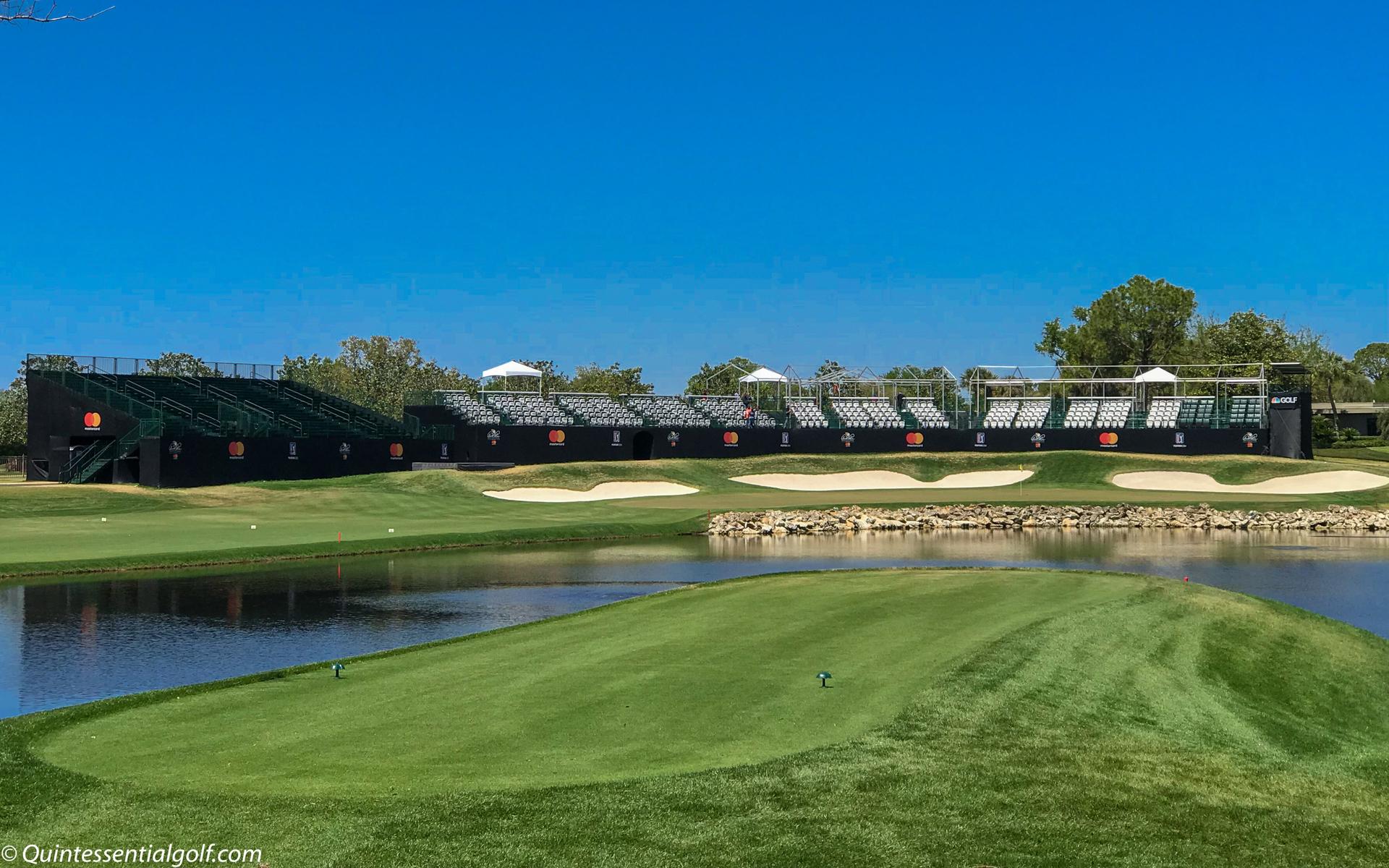 A island tee box surrounded by a water hazard with spectator stands towering over sand bunkers