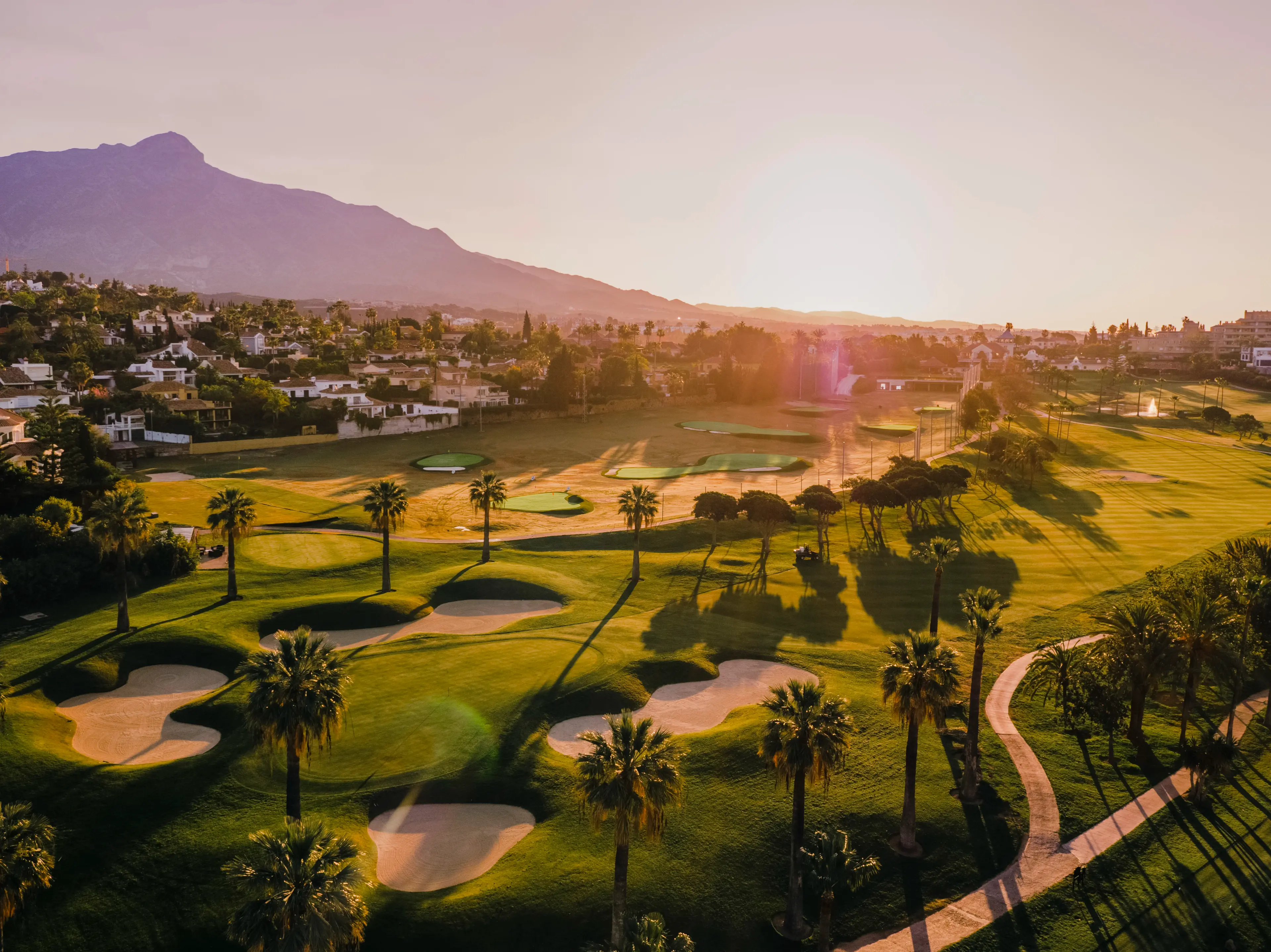 Overhead view of the sun setting over a well maintained fairway leading to a smooth green surrounded by sand bunkers
